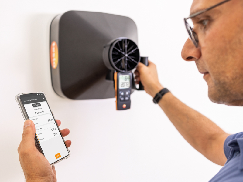 man holding a square thermal imaging instrument against a white wall while reading results on his phone app